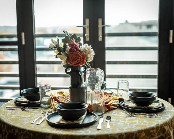 A table is set with gold blockprint table cloth, black ceramic bowls and plates, and a black ceramic vase full of felt flowers. 